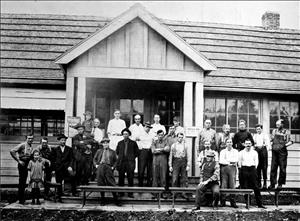 Large group of men in work clothes stands in front of a one story wooden building. One is holding a smiling child. Another child off to one side is frowning. 